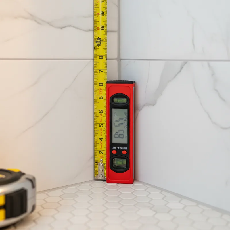 Close-up detail shot of a professional measuring tape against tile edge in a bathroom corner, with a digital level showing...
