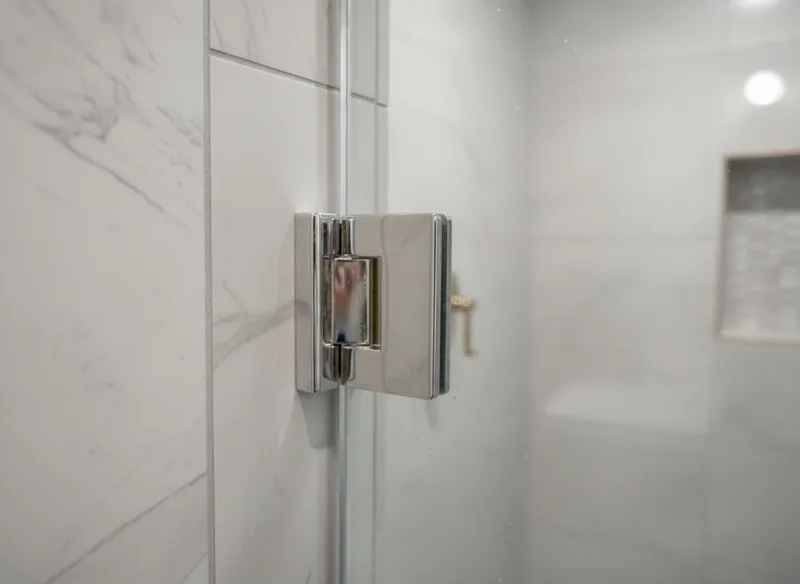 Close-up detail shot of premium chrome hinges and glass-to-wall brackets on a frameless shower door, showing polished meta...