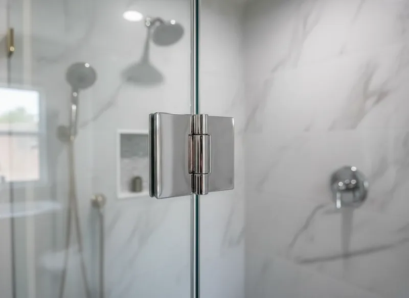 Close-up detail of a frameless glass shower door hinge and hardware mounted on clear tempered glass, with bright studio li...