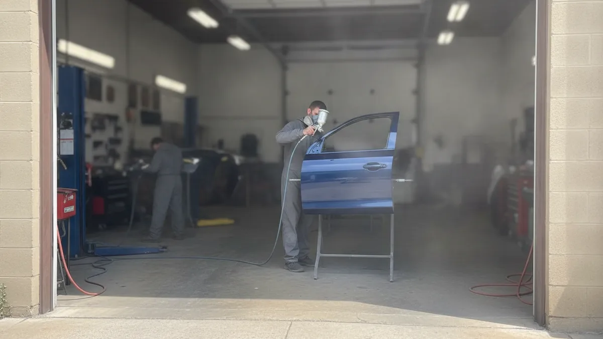 Auto technician spray-painting a vehicle panel in a professional body shop bay with natural daylight.