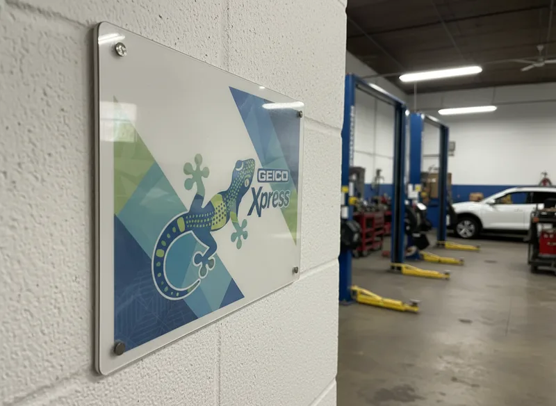 Close-up of a Geico Xpress certification badge or document displayed on a shop wall, with a professional repair facility e...