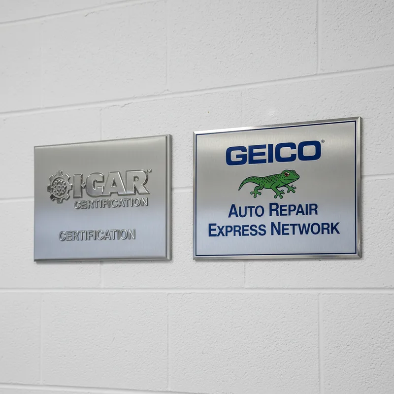 A close-up detail shot of an I-CAR and Geico certification badge or plaque mounted on a professional auto body shop wall, ...
